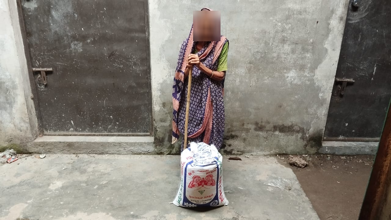 Elderly woman leaning on a walking stick beside a sack of rice